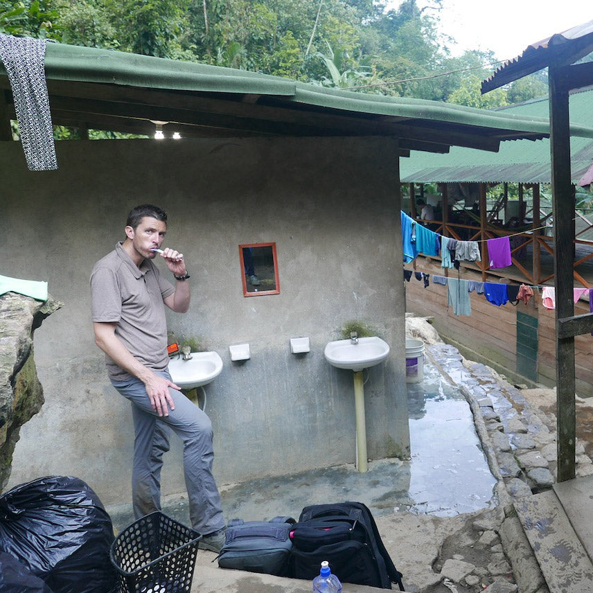A man brushes his teeth at an outdoor sink near a rustic building with laundry hanging in the background.