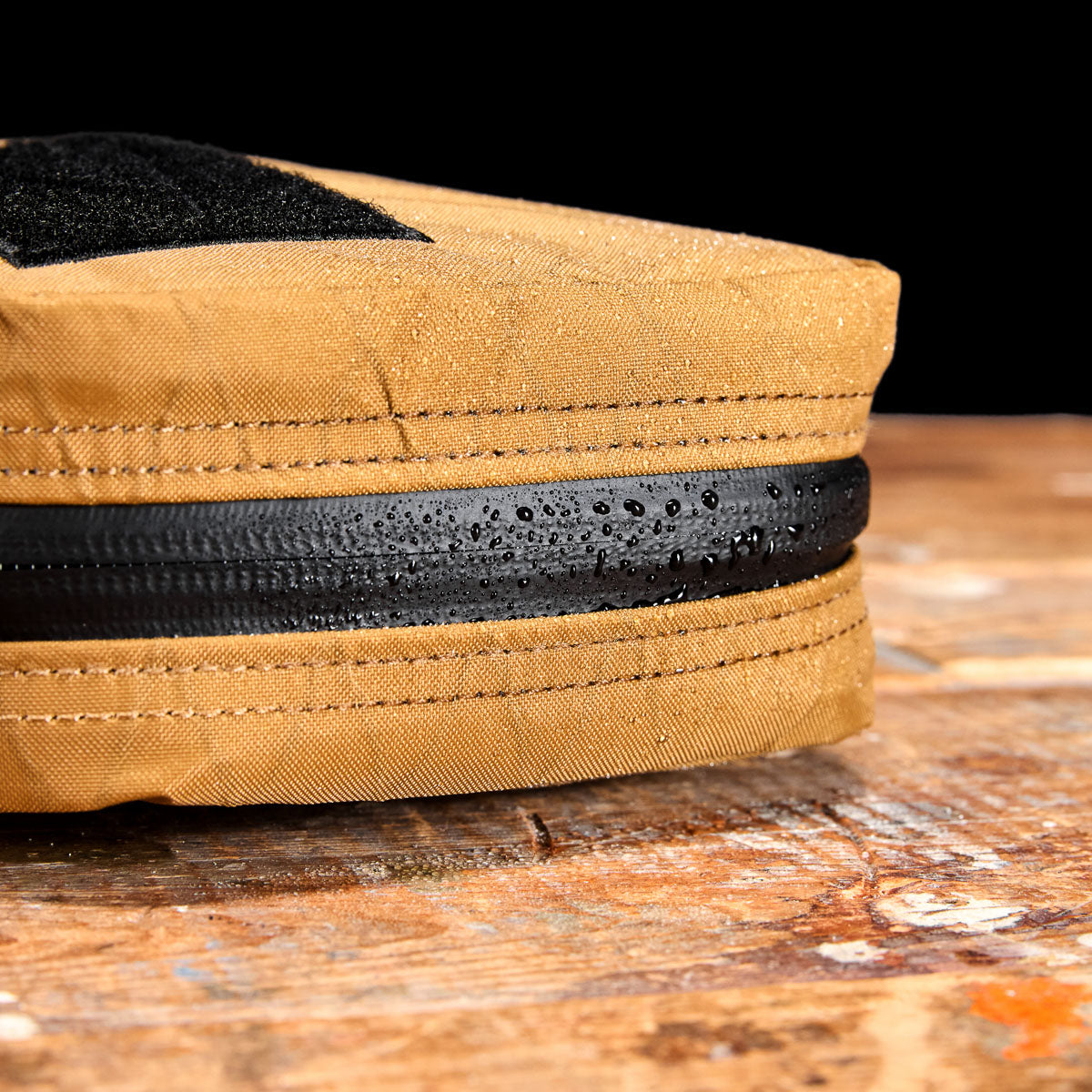 Close-up of a tan fabric pouch with water droplets on a black zipper, resting on a wooden surface.
