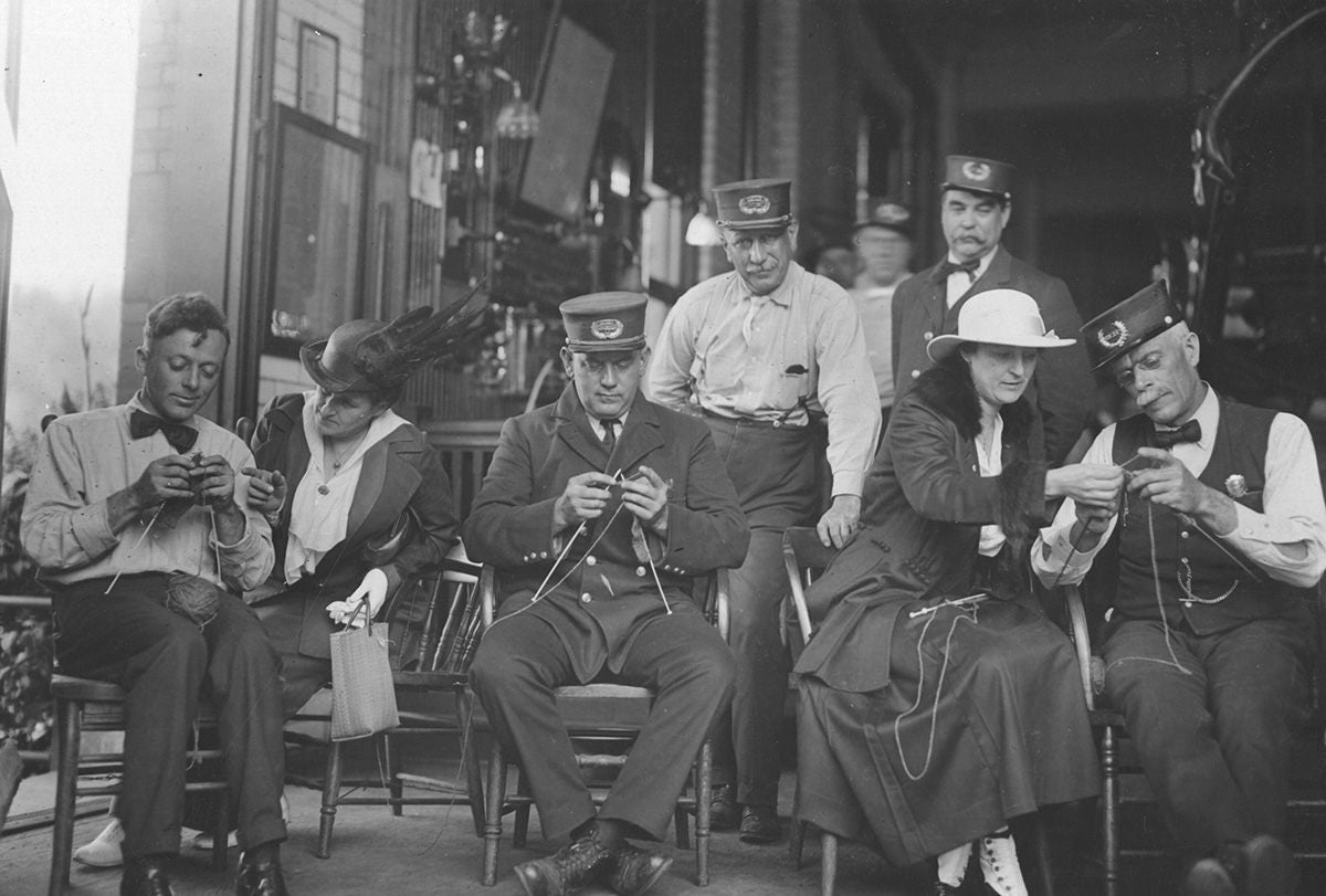 A group of men and women in vintage clothing sit knitting together, some wearing hats and uniforms.