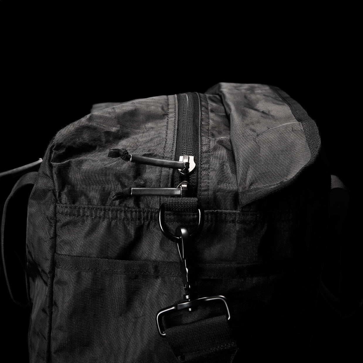 Close-up of a black nylon bag with a zipper and metal clip, set against a black background.