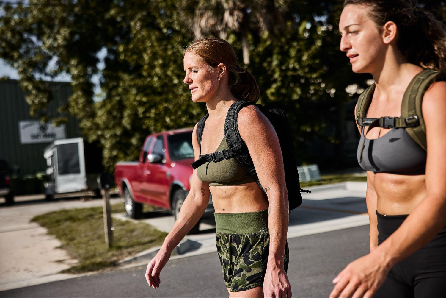 Two women in sports bras and shorts walk outdoors with backpacks on a sunny day.