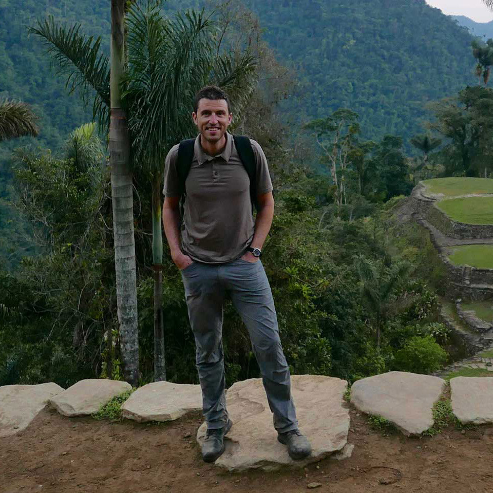 Man in hiking clothes stands on a stone path with lush green mountains and trees in the background.