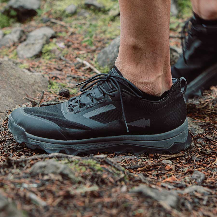 Close-up of a person wearing black trail running shoes on a forest path with rocks and moss.