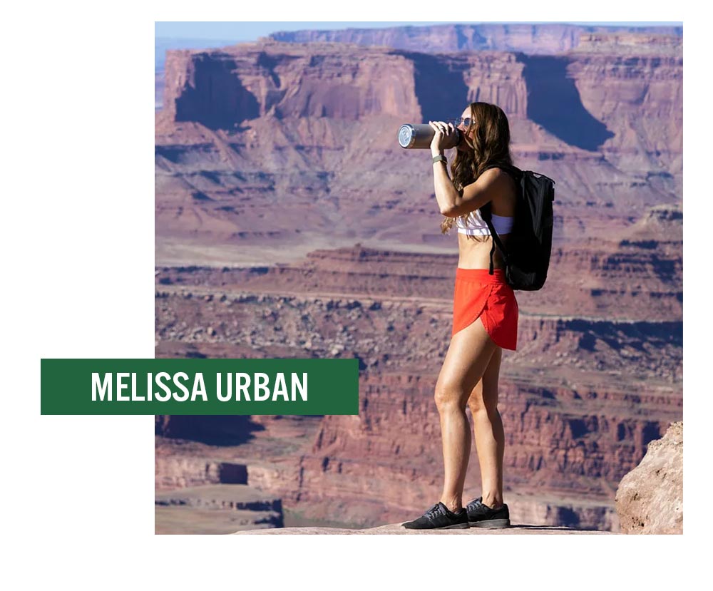 Woman in athletic wear drinks from a bottle while standing on a canyon cliff, with