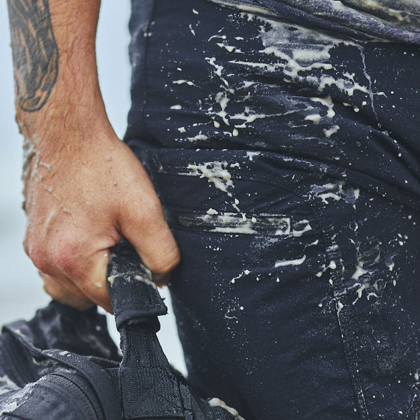 Close-up of a person's hand holding a bag, with sand and salt stains on their dark pants.