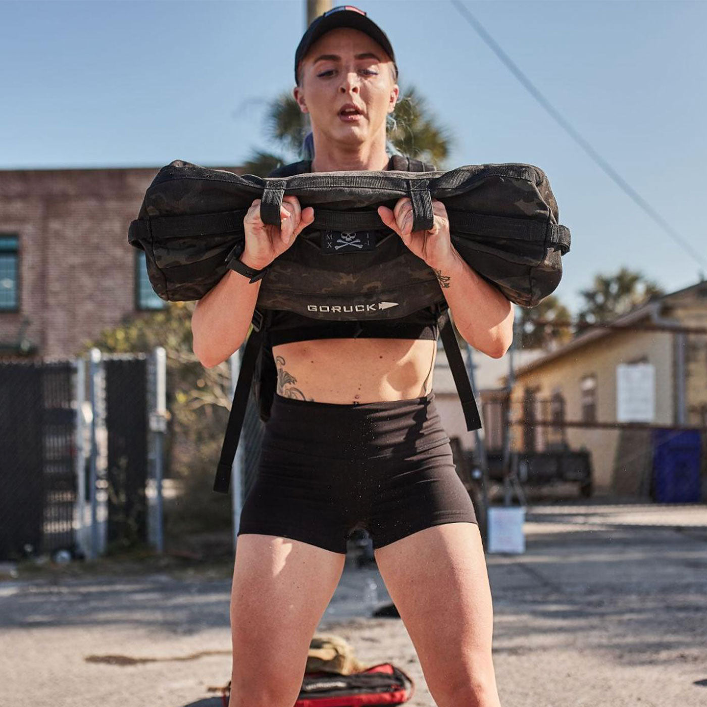 Person in athletic wear lifting a sandbag outside on a sunny day, with buildings and a fence in the background.
