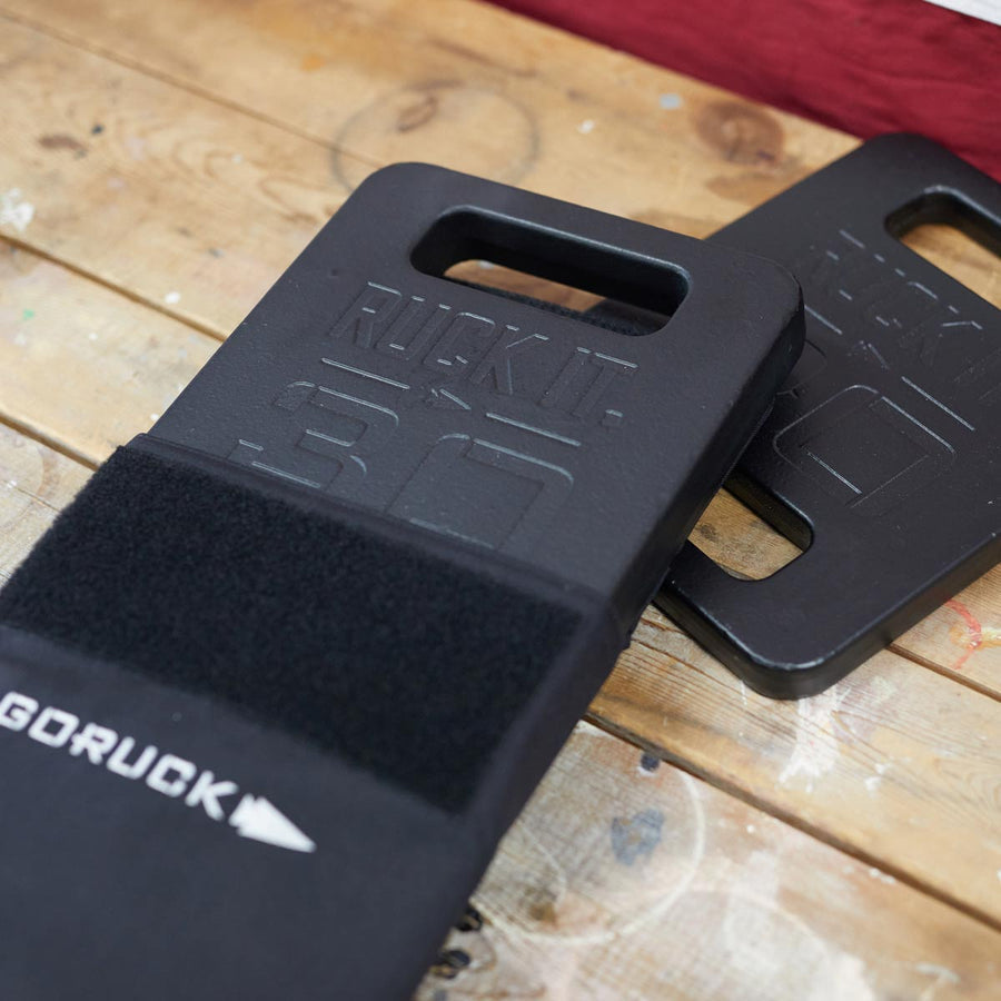 Two black GORUCK 30-pound ruck plates on a wooden surface, one stacked partly over the other.