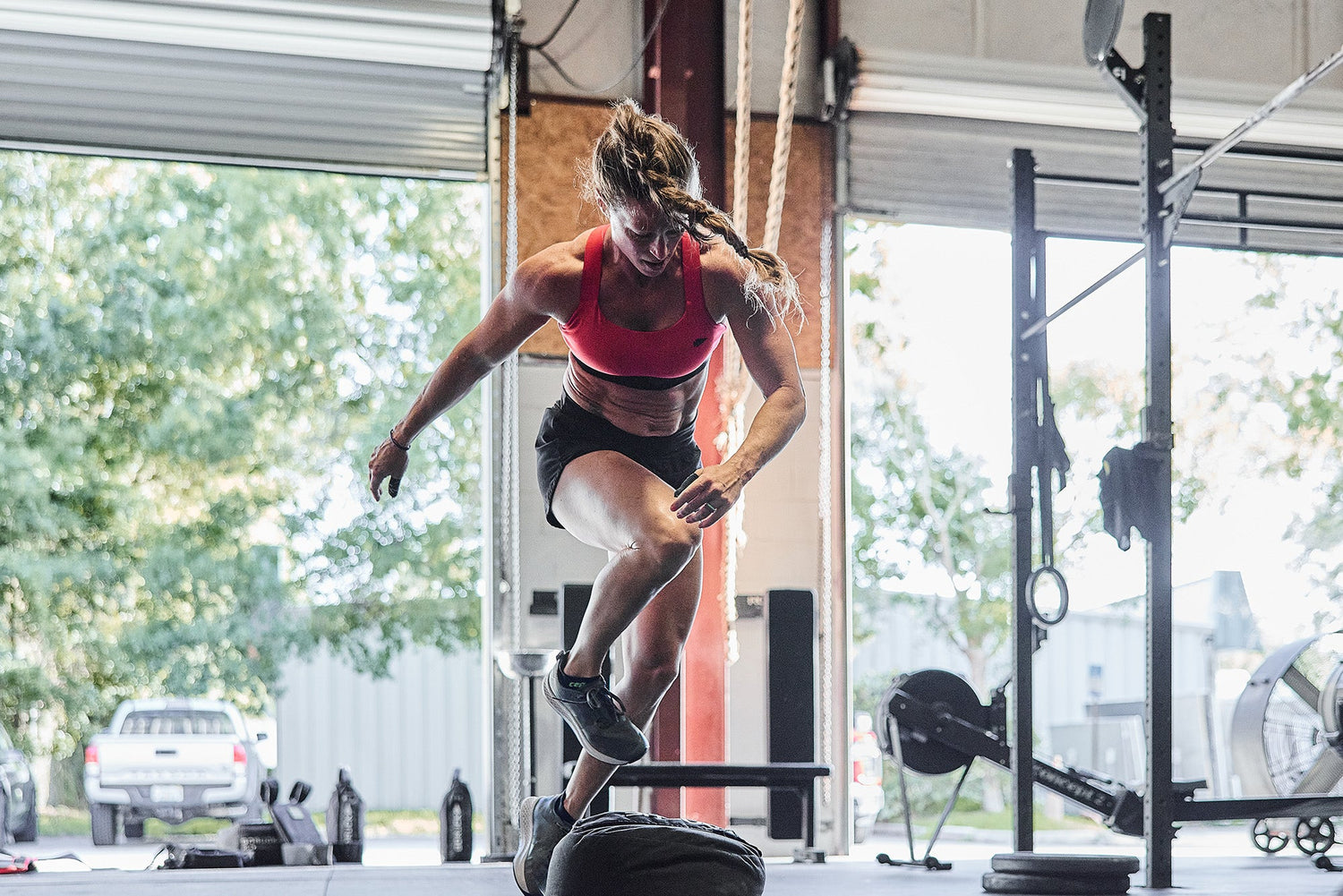 Woman in athletic wear jumps onto a tire during a workout in a gym with open garage doors.