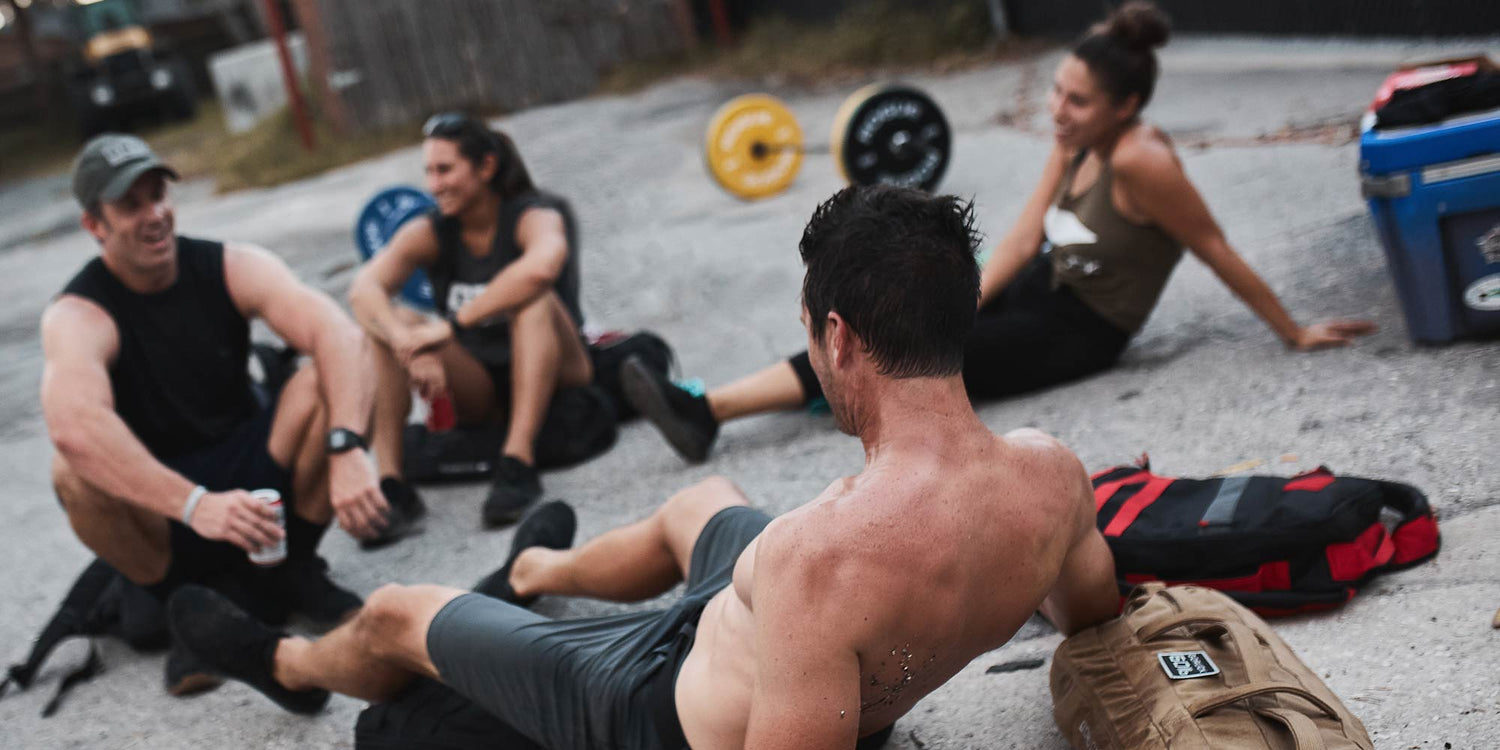 Four people in workout clothes relax and chat outdoors on a paved surface with gym weights in the background.