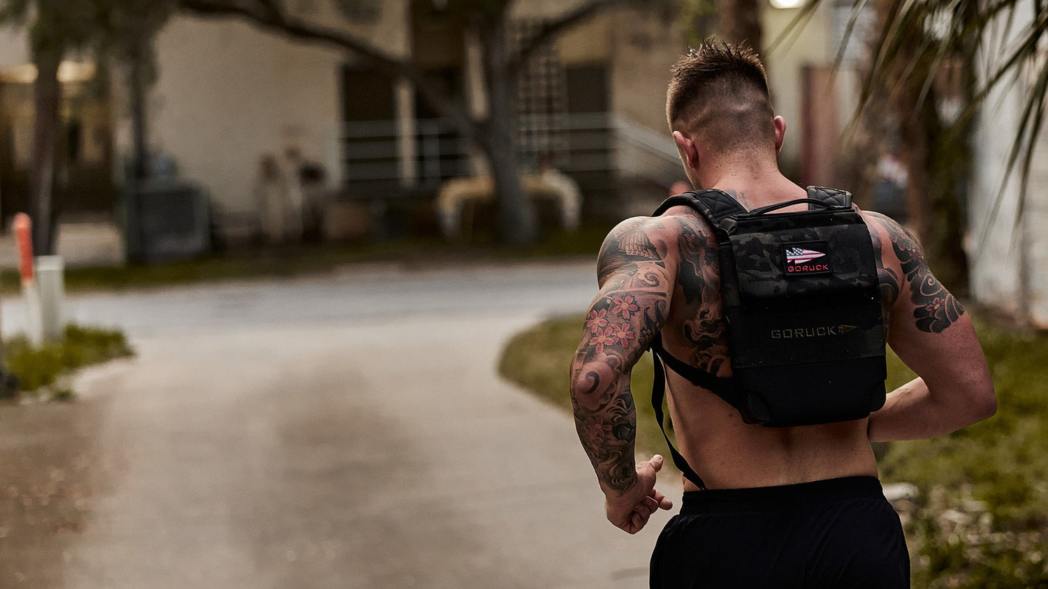 Tattooed person runs outdoors, wearing a black weighted vest and athletic shorts on a paved path.