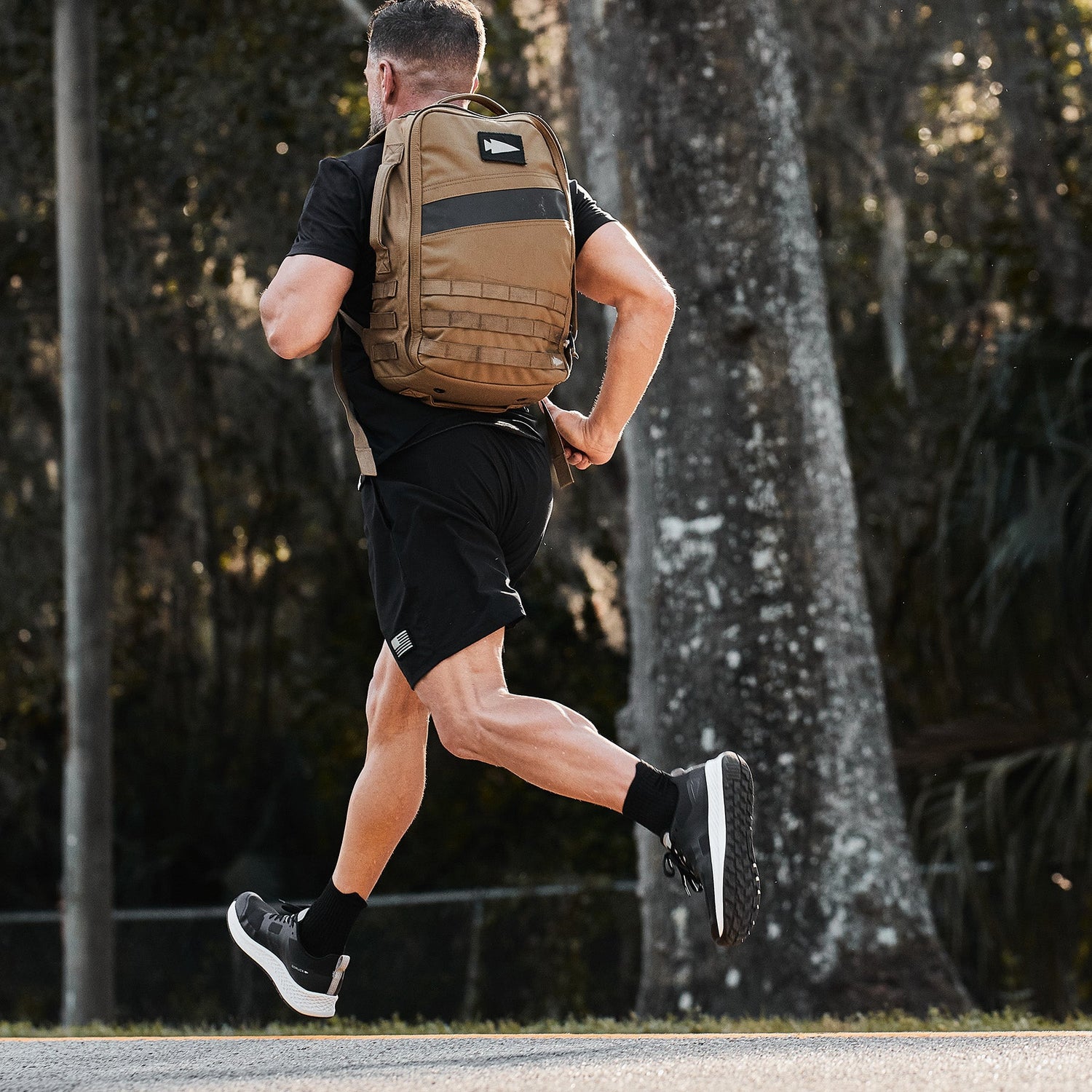Man jogging outdoors wearing black athletic gear and a brown backpack, with trees in the background.