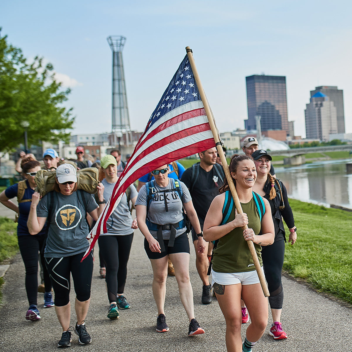 Group of people walking outdoors by a river; one woman in front carries a large American flag and smiles.