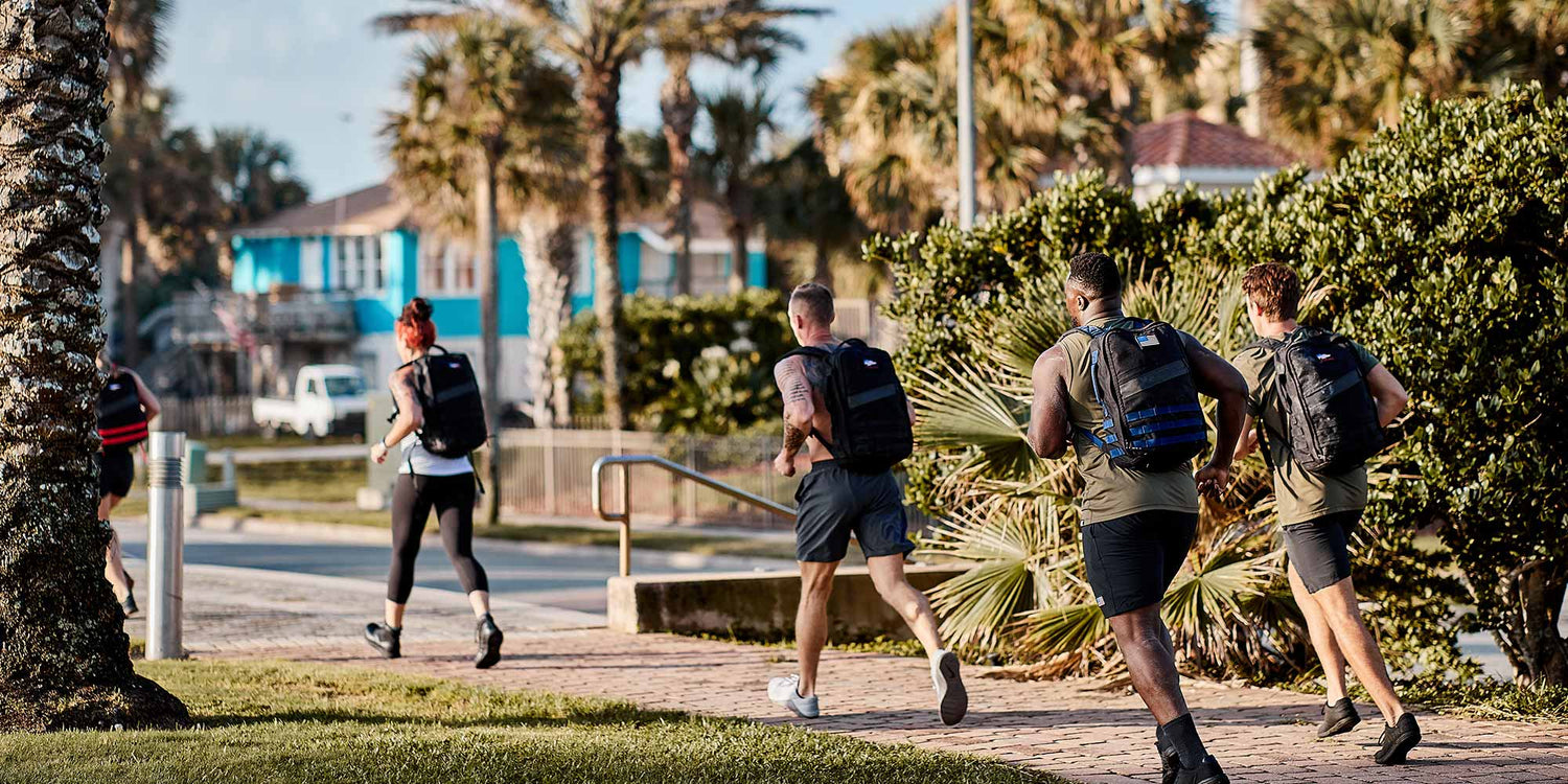 Five people with backpacks run on a sunny sidewalk lined with palm trees and houses in the background.