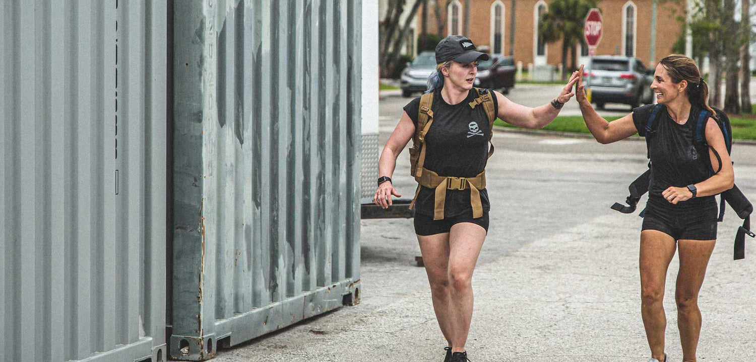 Two women in athletic gear high-five each other while running outdoors near a large metal container.