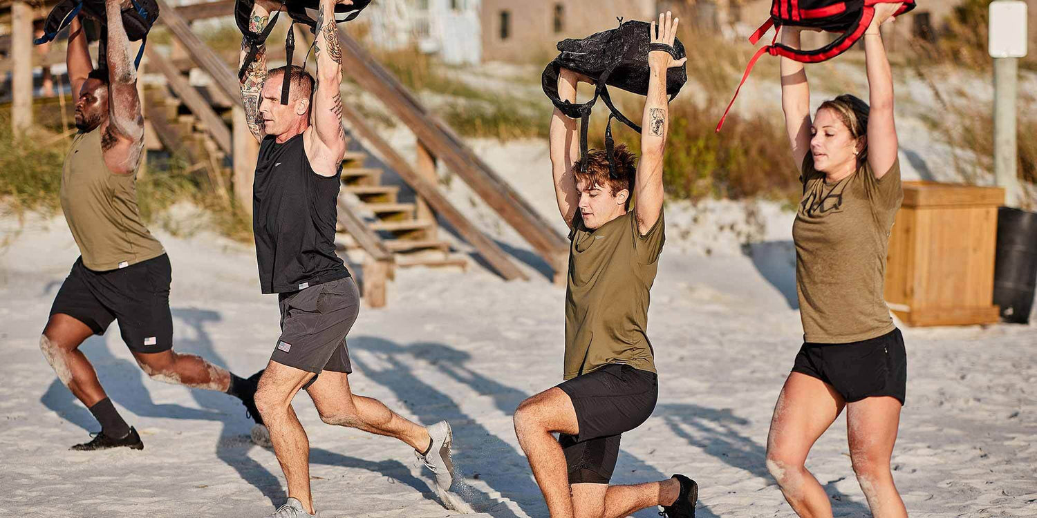 Four people doing lunges on the beach, lifting bags overhead during an outdoor workout.