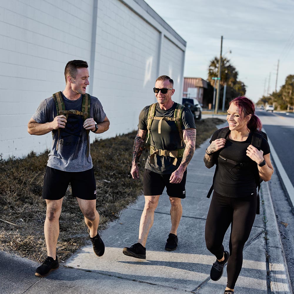 Three people with backpacks smile and walk together on a sidewalk by a street and white building.