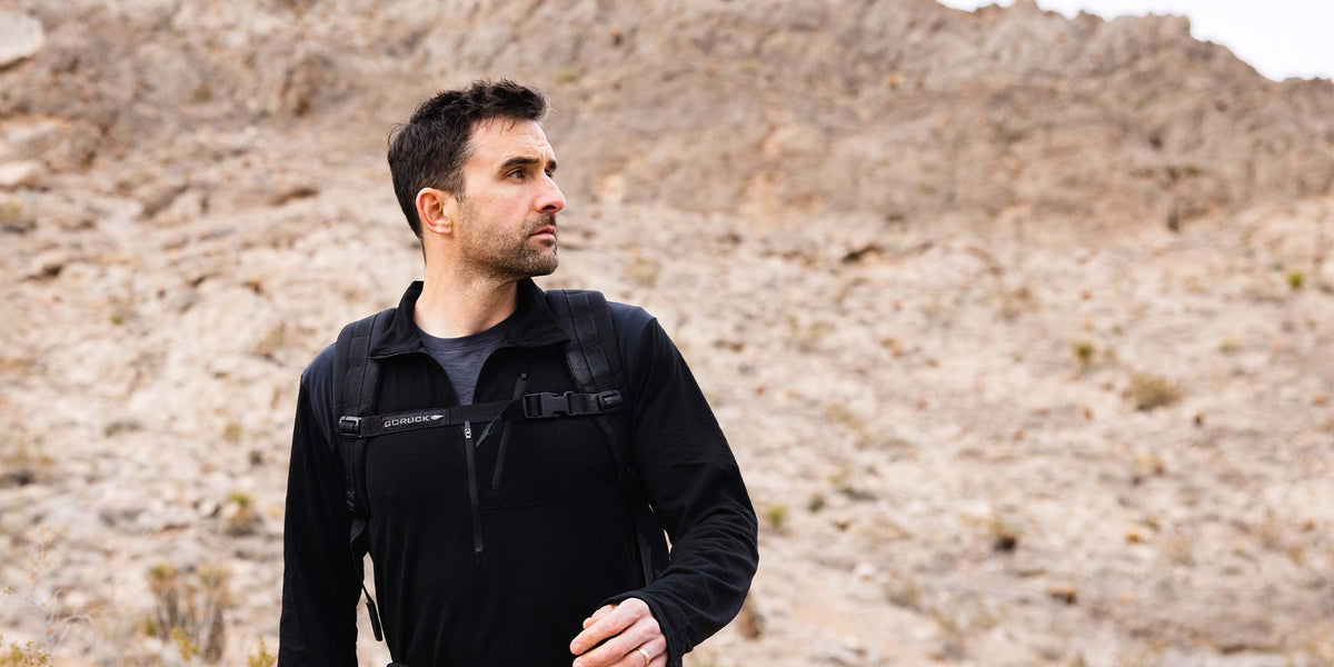 Man in a black jacket hiking in a rocky, desert landscape, looking off to the side.