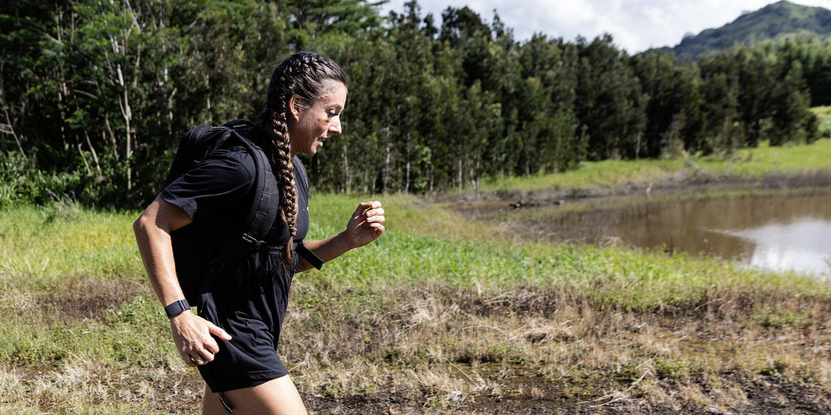 Woman with braided hair runs outdoors on a dirt trail near a pond, surrounded by grass and trees.
