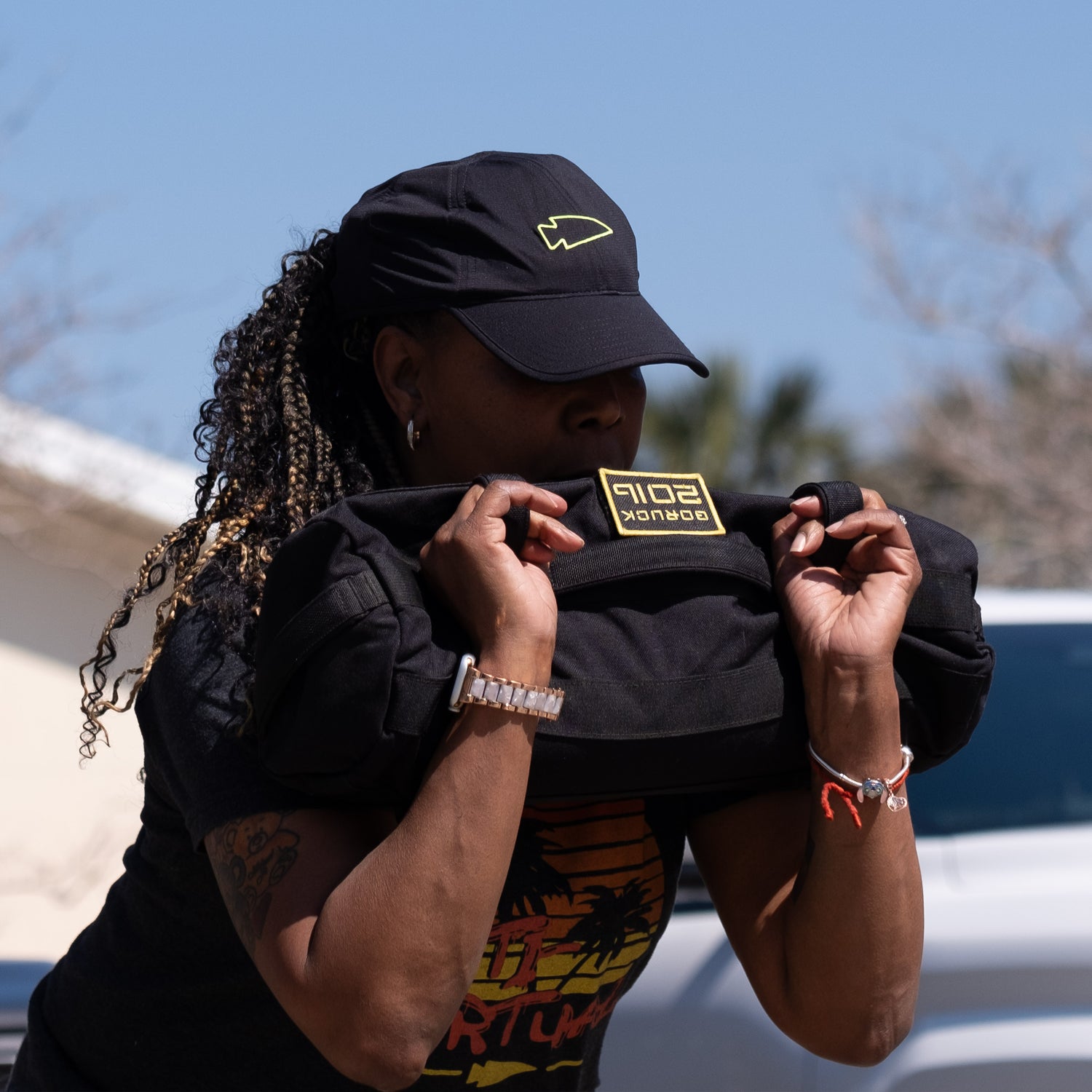 A woman lifts a weighted sandbag outdoors, wearing a black cap and t-shirt with a colorful graphic.