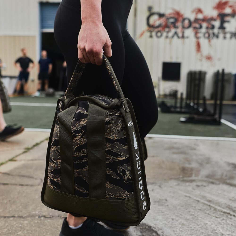 Person carrying a camouflage GORUCK bag outside a gym with people and equipment in the background.