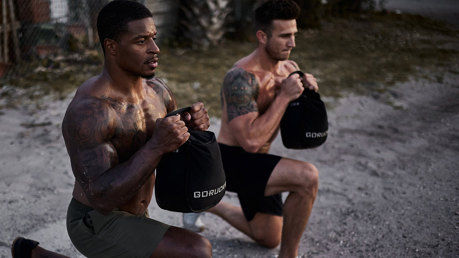 Two men with tattoos do weighted lunges outdoors, holding sandbags and wearing athletic shorts.