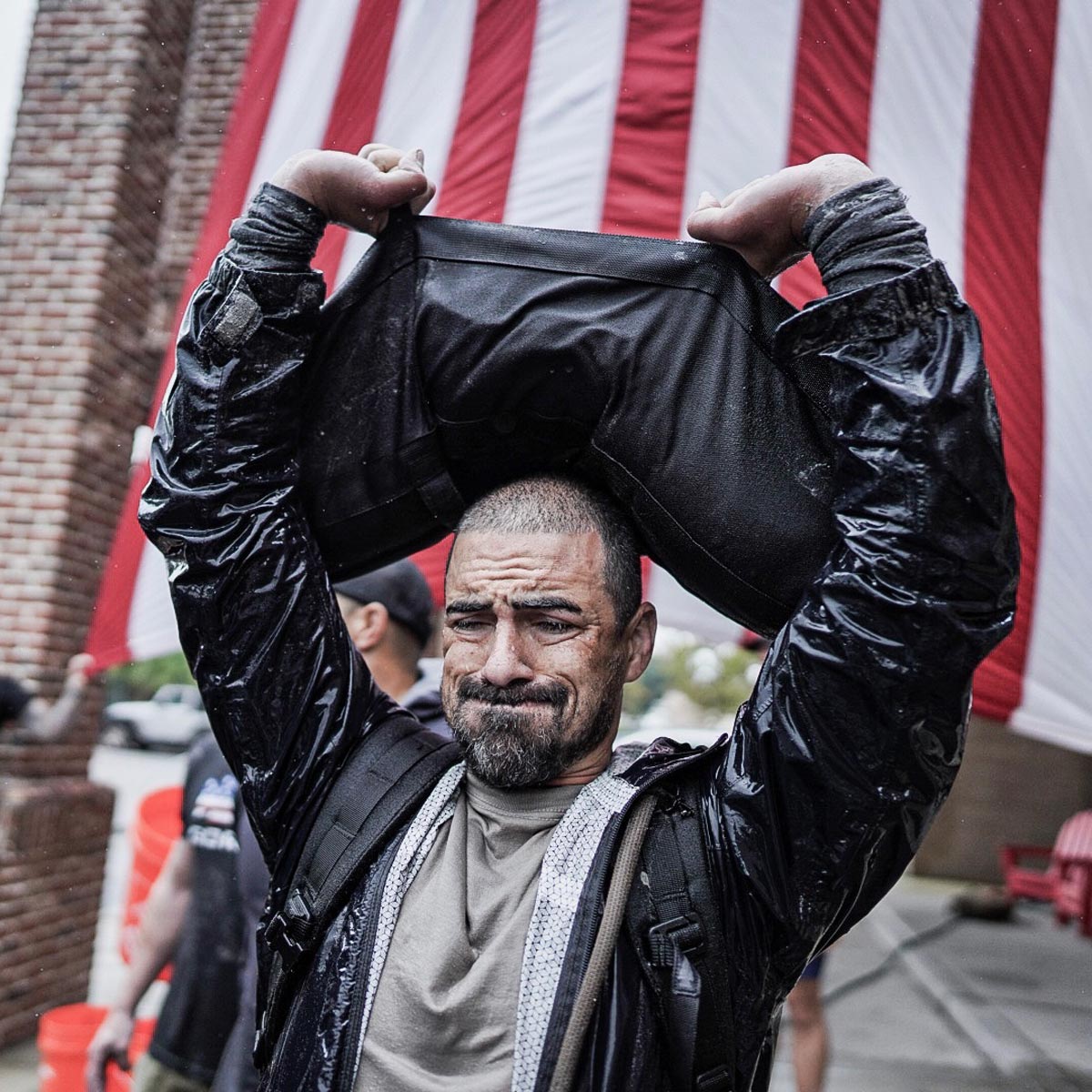 Man lifts heavy sandbag overhead, wearing a rain jacket, with a large American flag in the background.