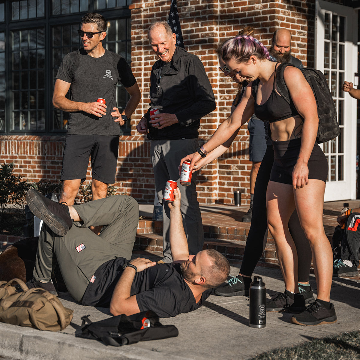 Five people in workout gear smiling and sharing drinks outside a brick building; one person lies on the ground.
