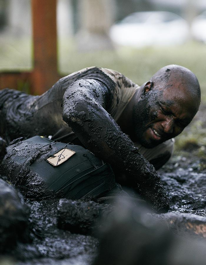 Man crawling through thick mud with a strained expression, covered in dirt, participating in an outdoor challenge.