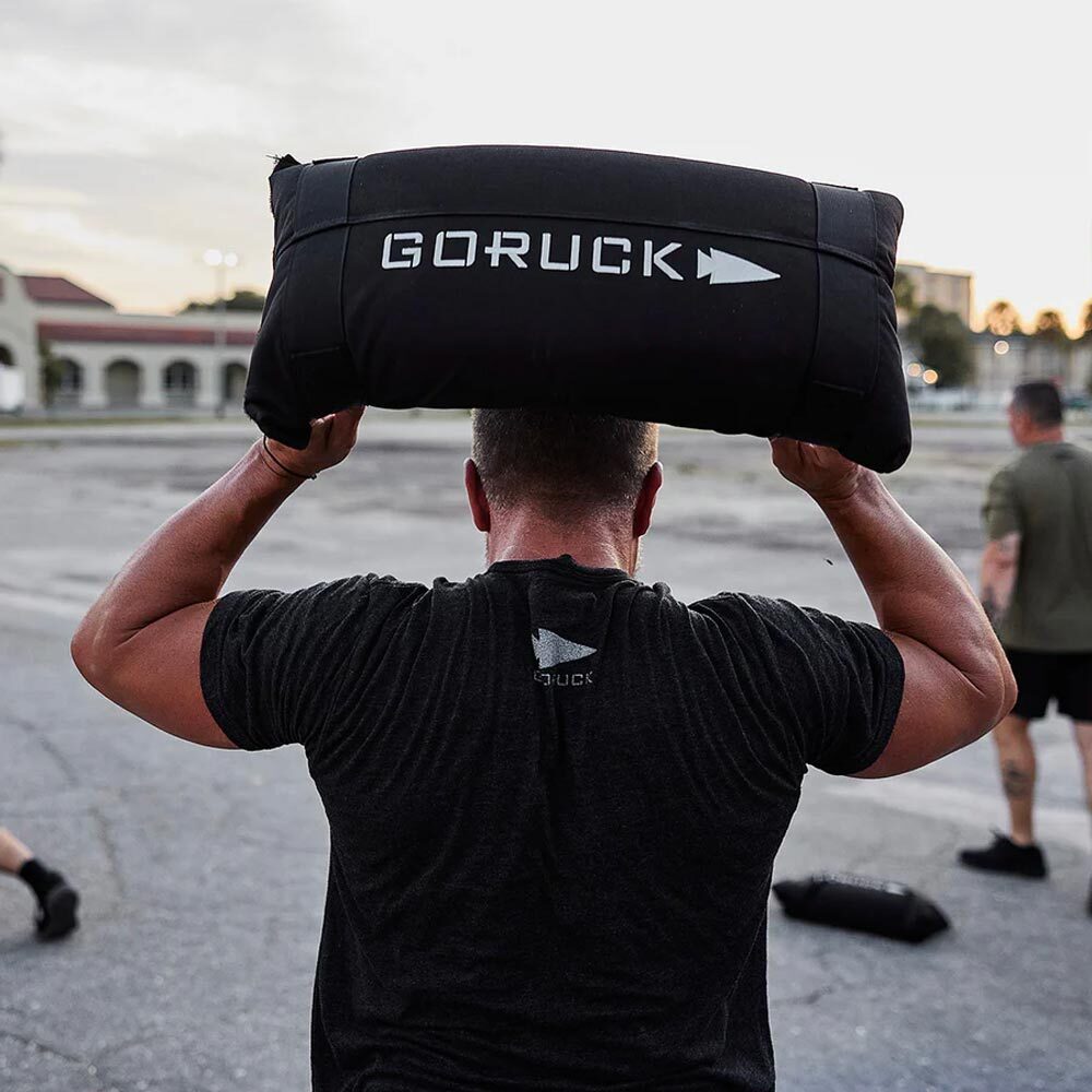 Person lifting a GORUCK sandbag overhead outdoors, with others exercising in the background.