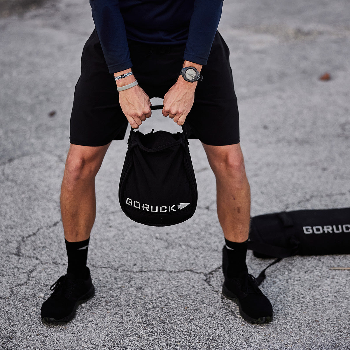 Person holding a GORUCK sandbag between their legs, standing on pavement in athletic wear.