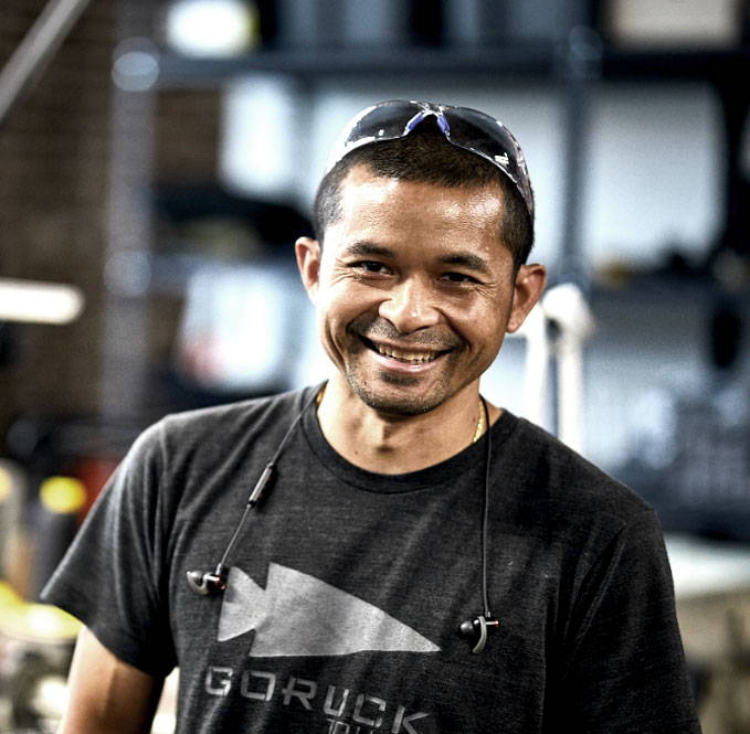 Smiling man wearing safety glasses and a GORUCK t-shirt stands indoors with headphones around his neck.