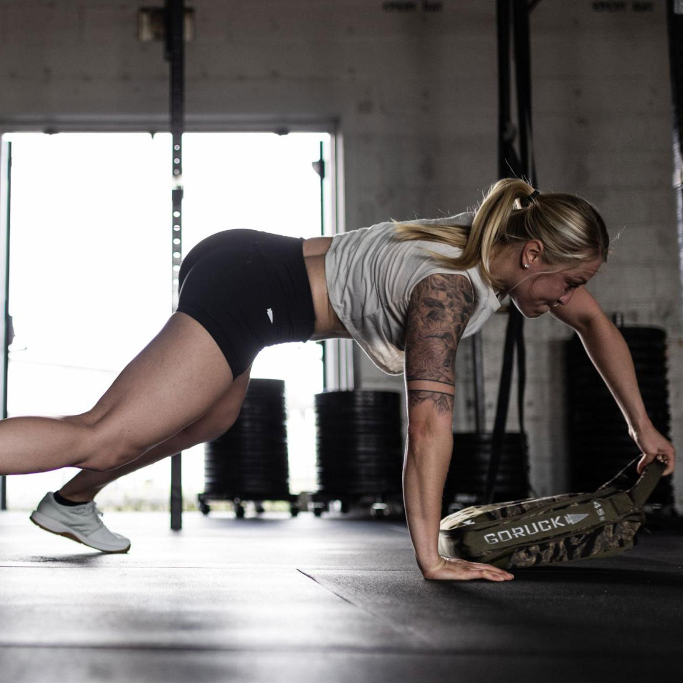 Woman doing a plank exercise in a gym, holding a weighted sandbag with one hand on the floor.