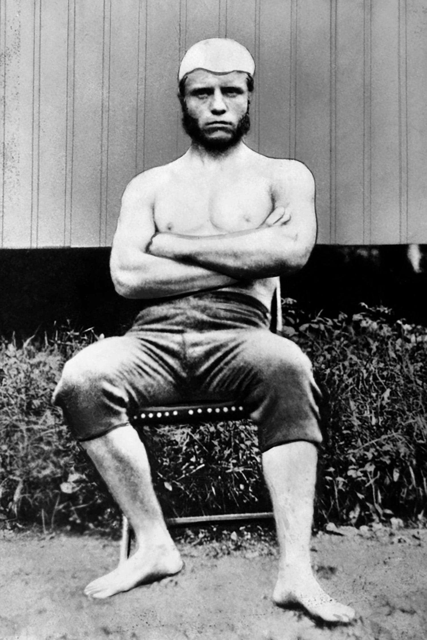Black-and-white photo of a shirtless man in a swim cap, sitting on a chair with arms crossed.