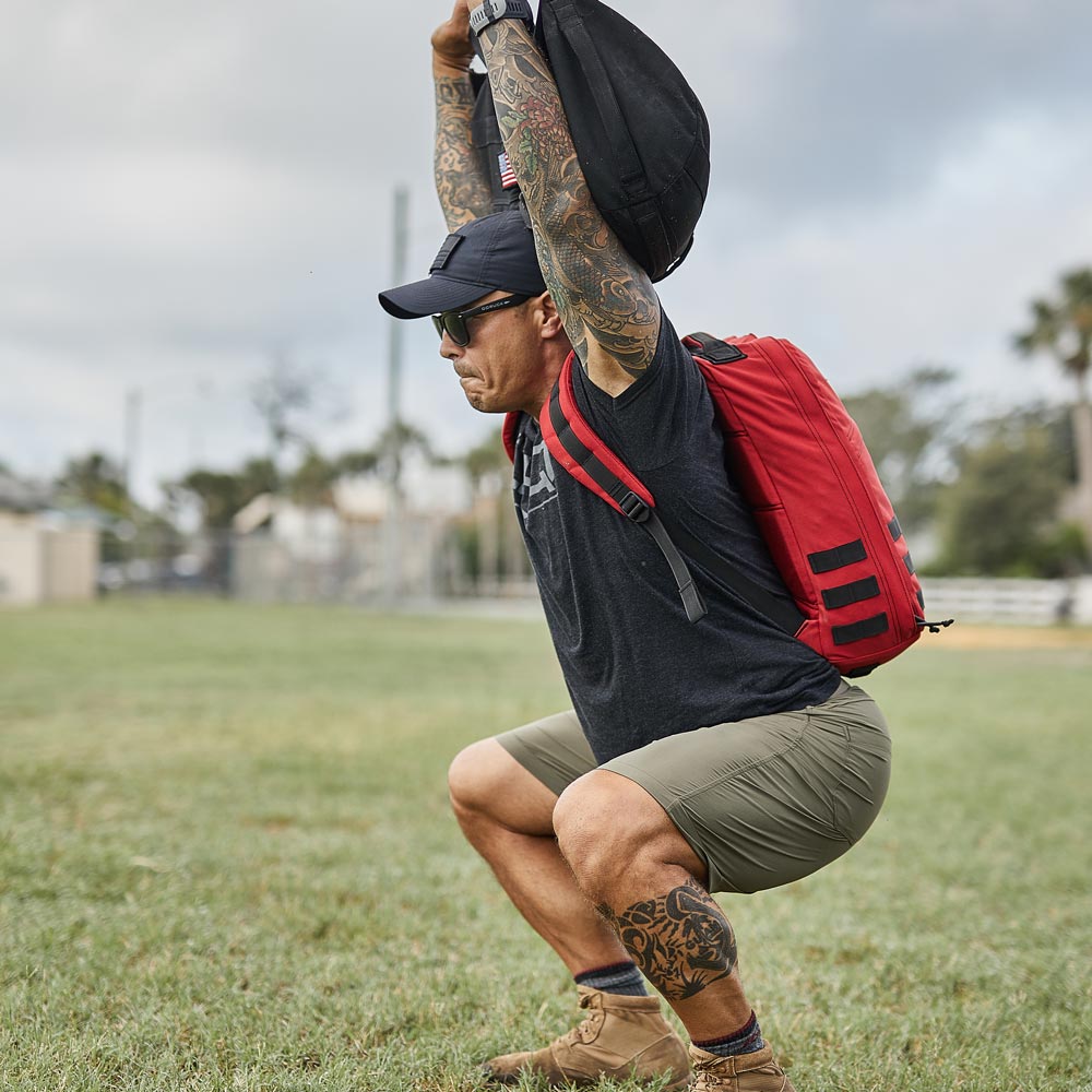 Man with tattoos squats outdoors, lifting a weighted bag overhead, wearing a red backpack and sunglasses.