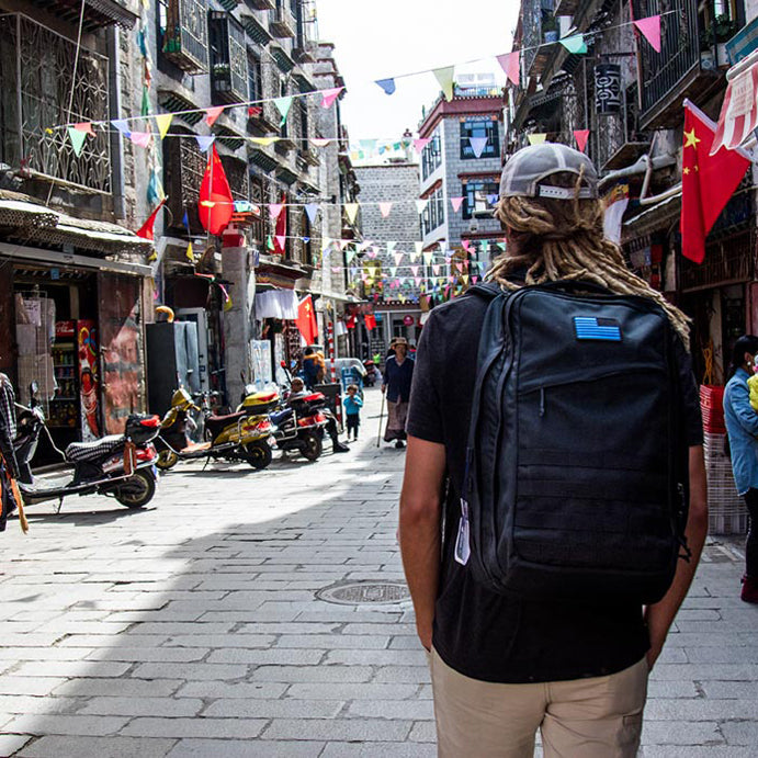 Person with backpack and dreadlocks walks down a lively, flag-decorated street with scooters and people.