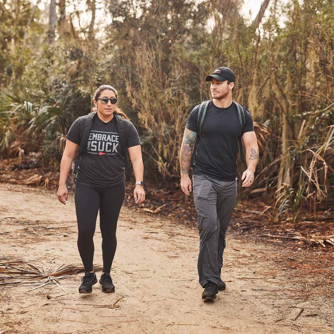 Two people in athletic clothing walk on a dirt trail through a wooded area, carrying backpacks.