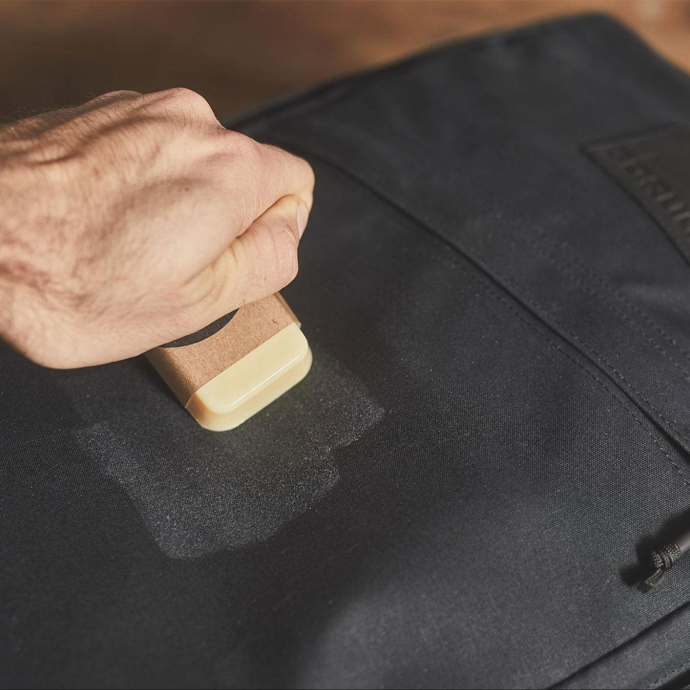 A hand applies wax to a black fabric bag using a rectangular wax block.
