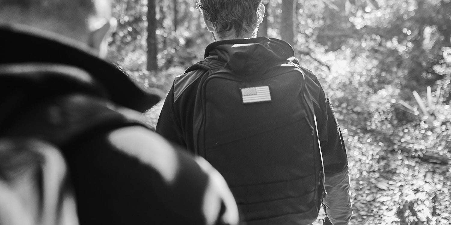Two people hiking in a forest, one wearing a backpack with an American flag patch, in black and white.