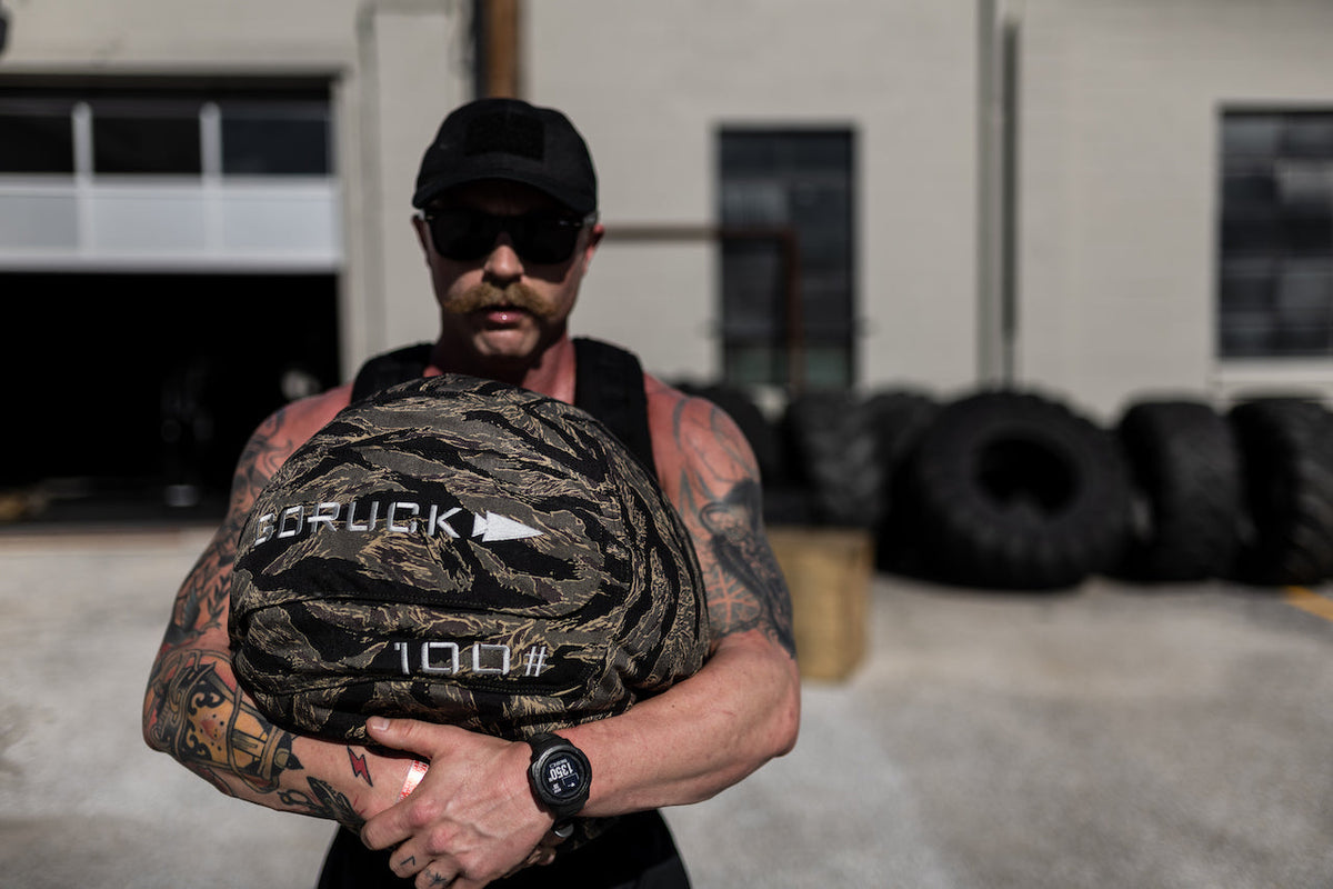 Man holding GORUCK 100lb sandbag outdoors, wearing fitness gear and sunglasses