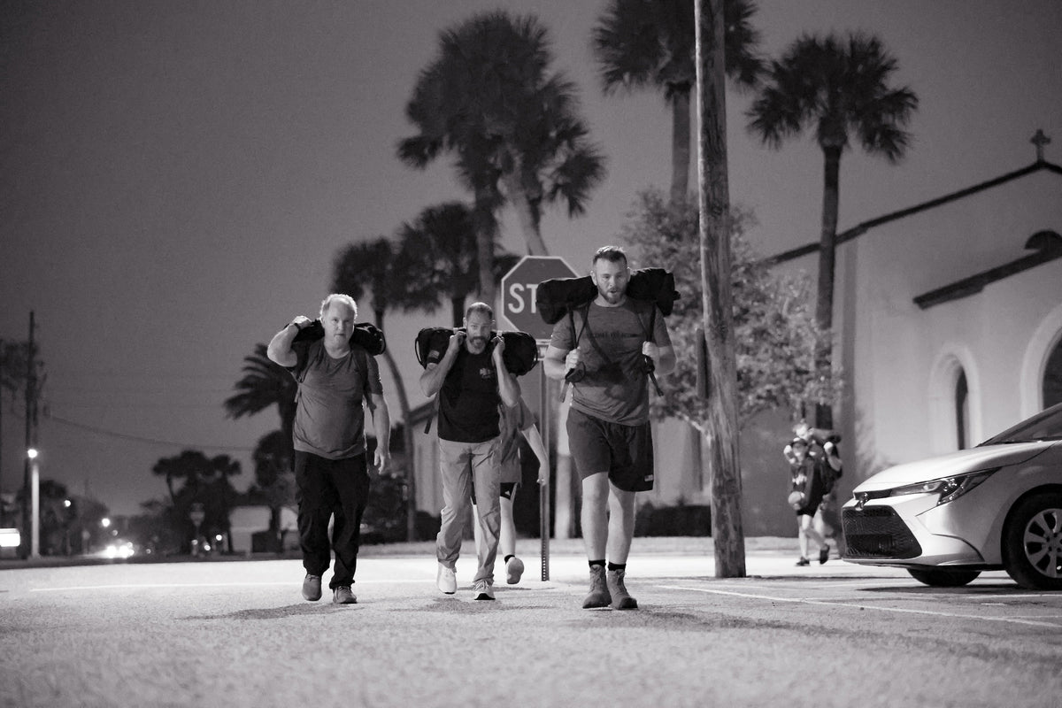 Three men rucking with sandbags on a city street at night, palm trees in background, GORUCK gear