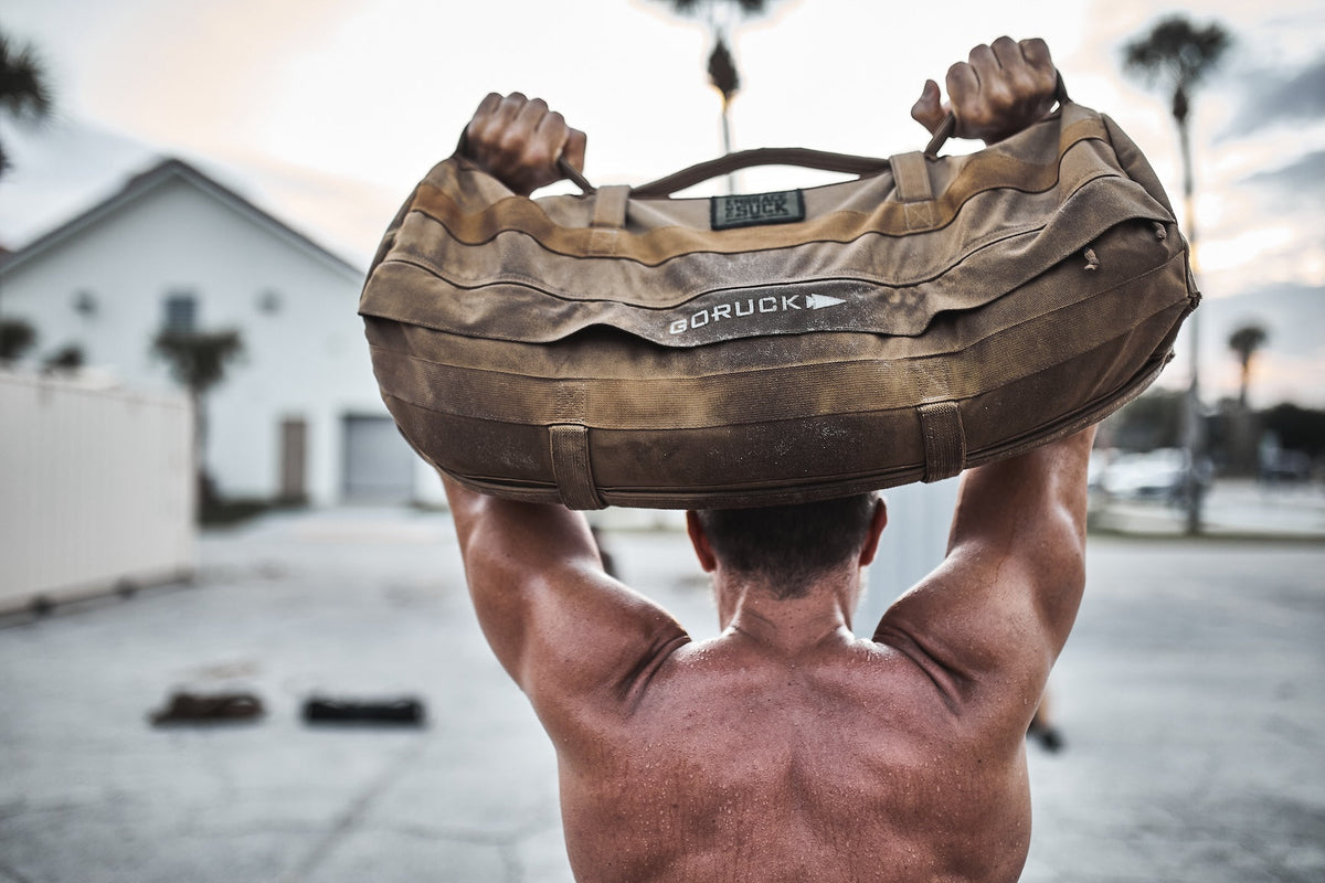 Muscular man lifts GORUCK rucking sandbag outdoors, showcasing durable fitness gear