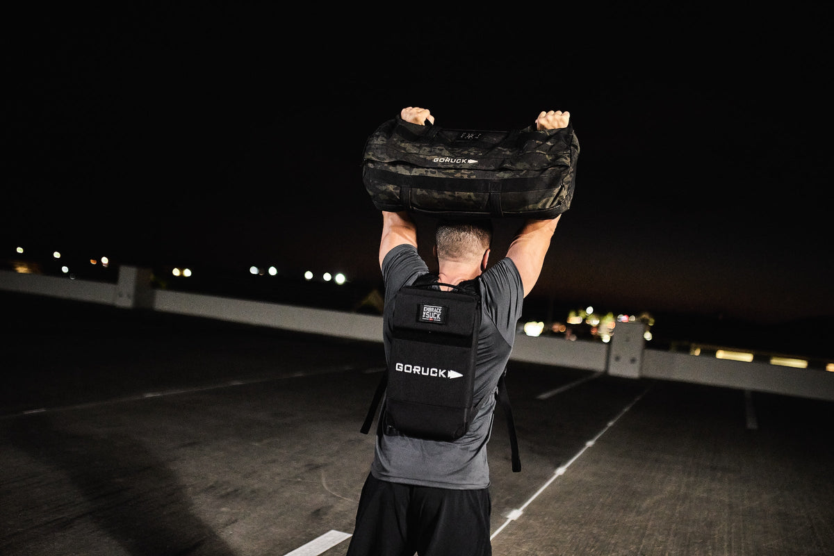 Man lifting GORUCK camo duffel on rooftop at night, wearing GORUCK rucksack, outdoor fitness