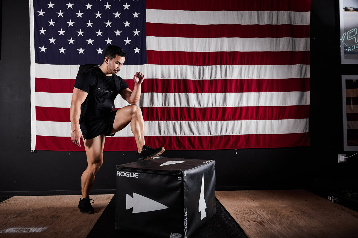 Man wearing GORUCK rucksack training in gym, stepping onto Rogue plyo box with American flag backdrop.