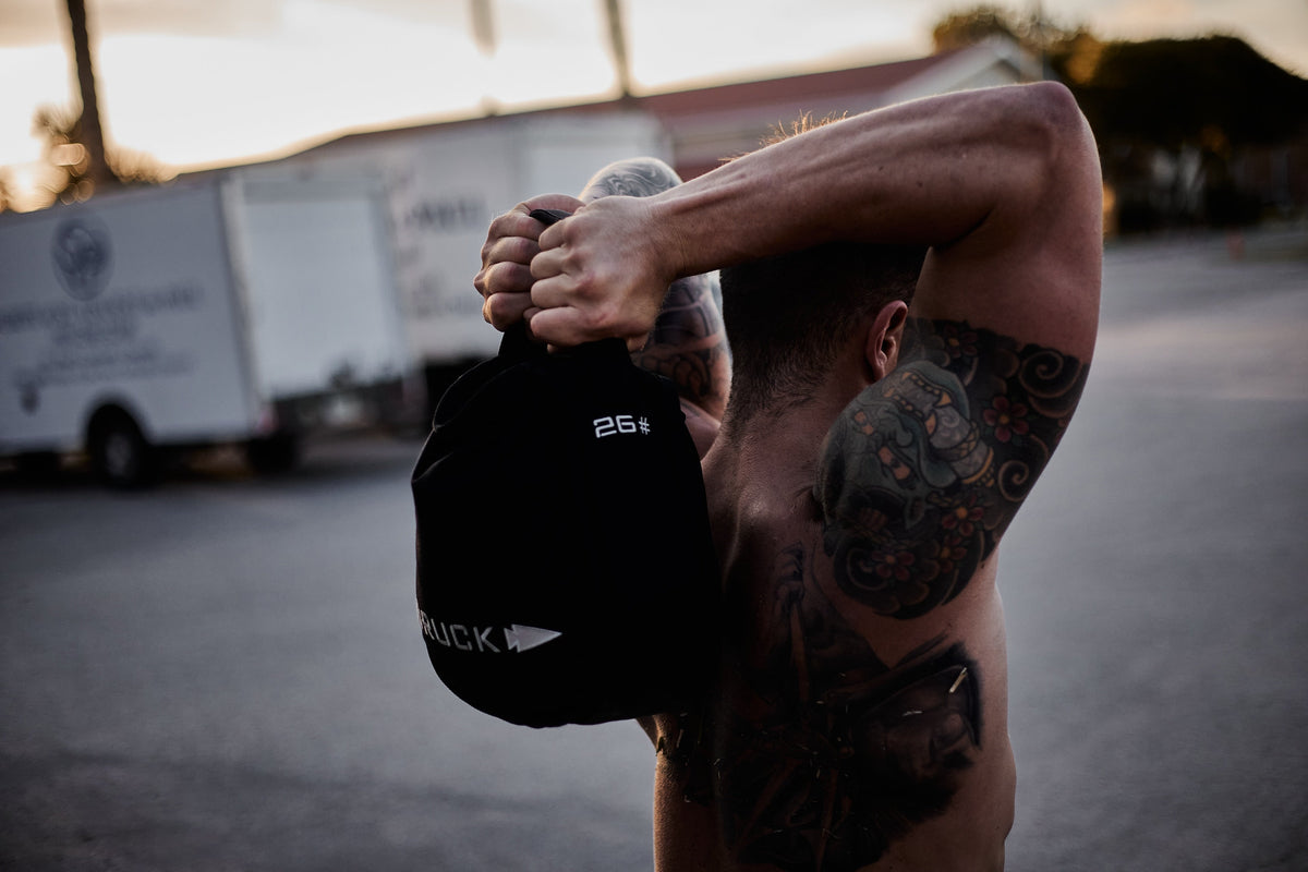 Tattooed man outdoors lifting GORUCK 26 lb sandbag, showcasing tough rucking gear