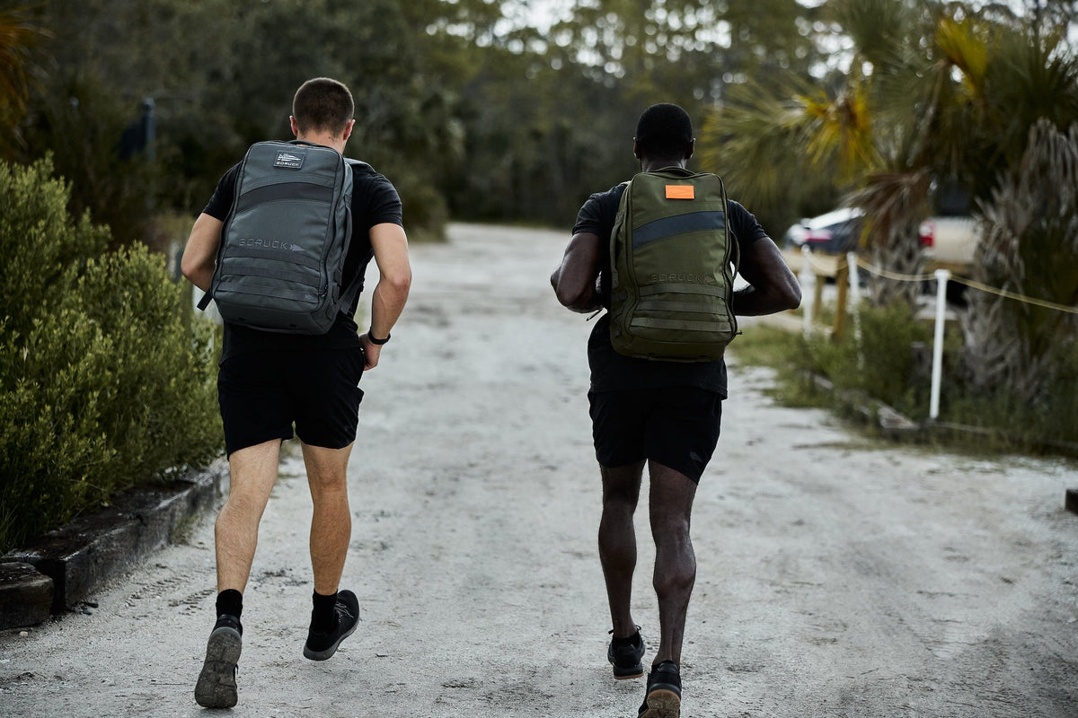 Two men wearing GORUCK backpacks rucking outdoors on a dirt path surrounded by greenery