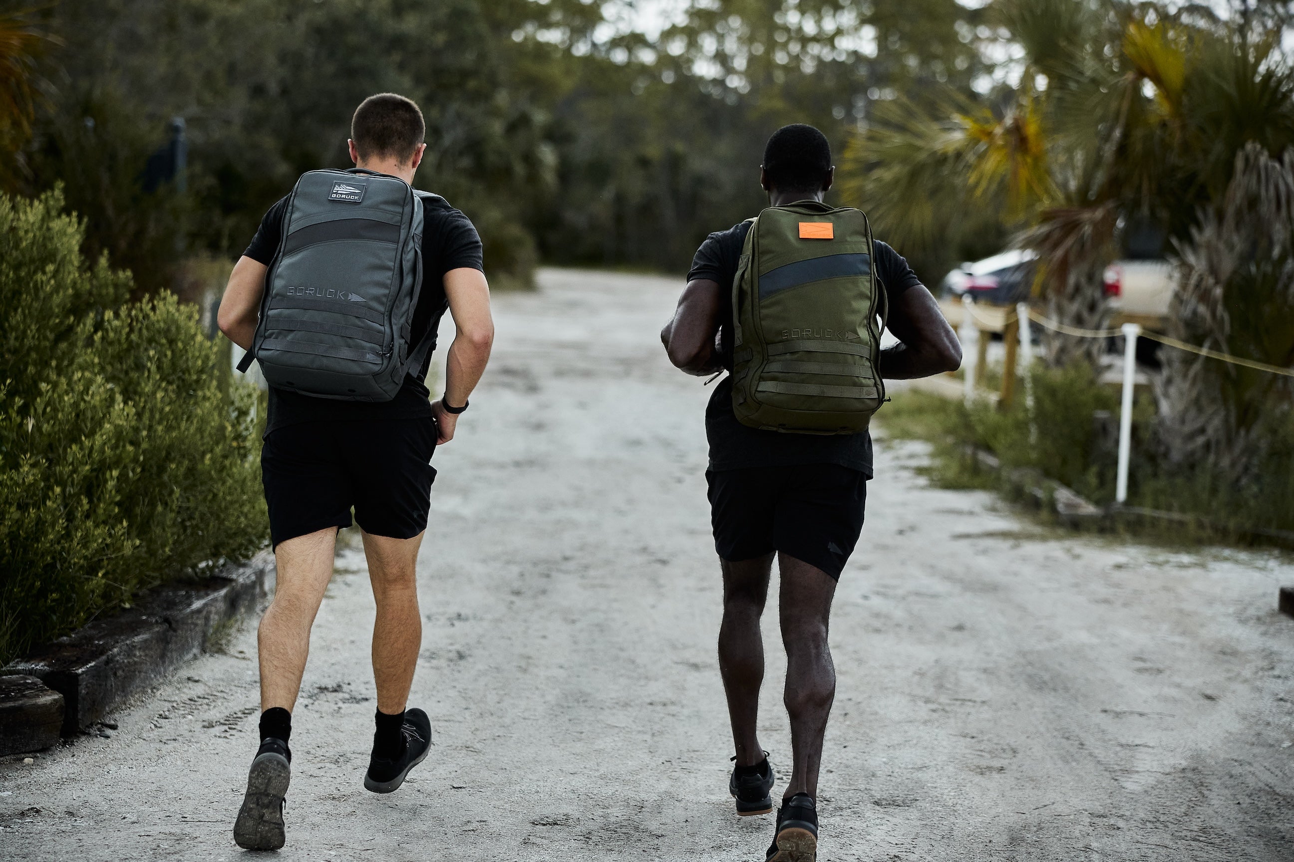Two men wearing GORUCK backpacks rucking outdoors on a dirt path surrounded by greenery