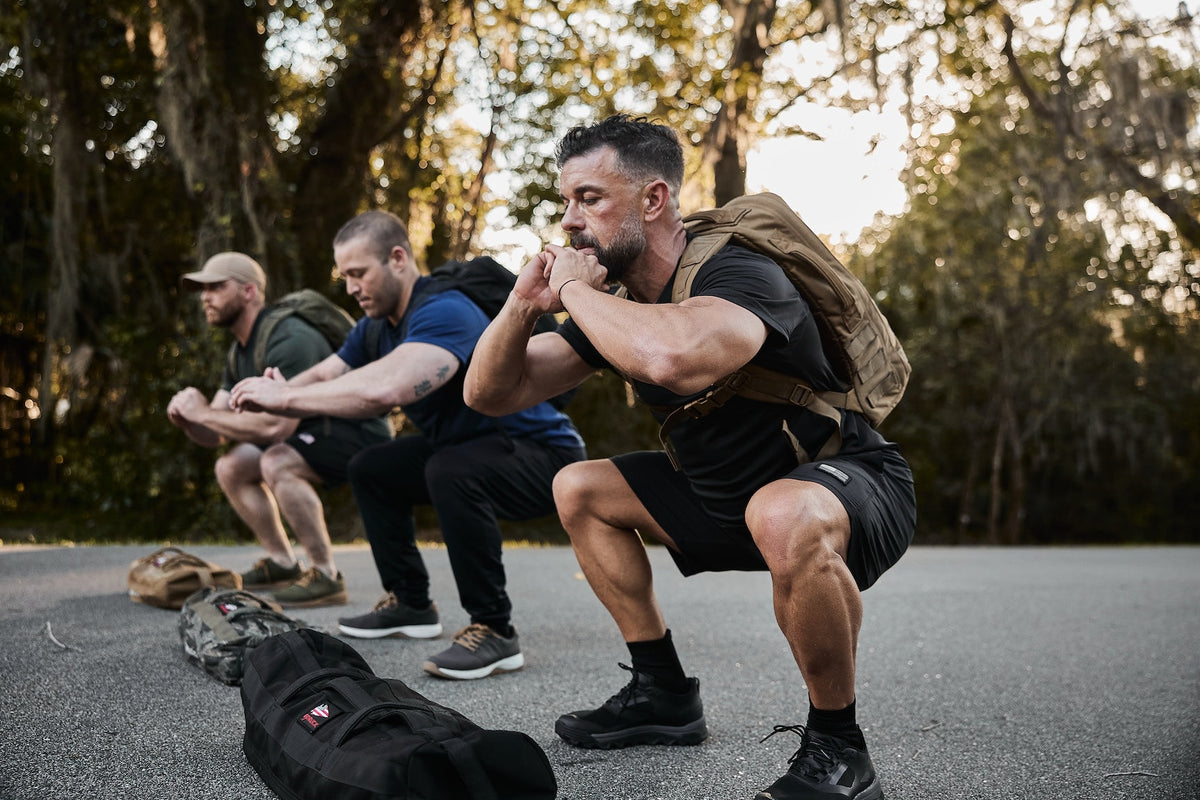 Three men wearing GORUCK rucksacks doing outdoor squats in workout gear on a paved path