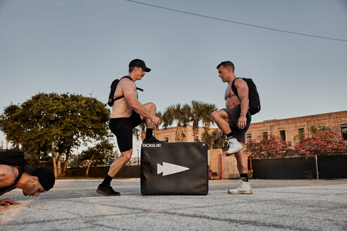 Two men wearing GORUCK rucksacks exercise outdoors with a Rogue plyo box, another man doing push-ups.