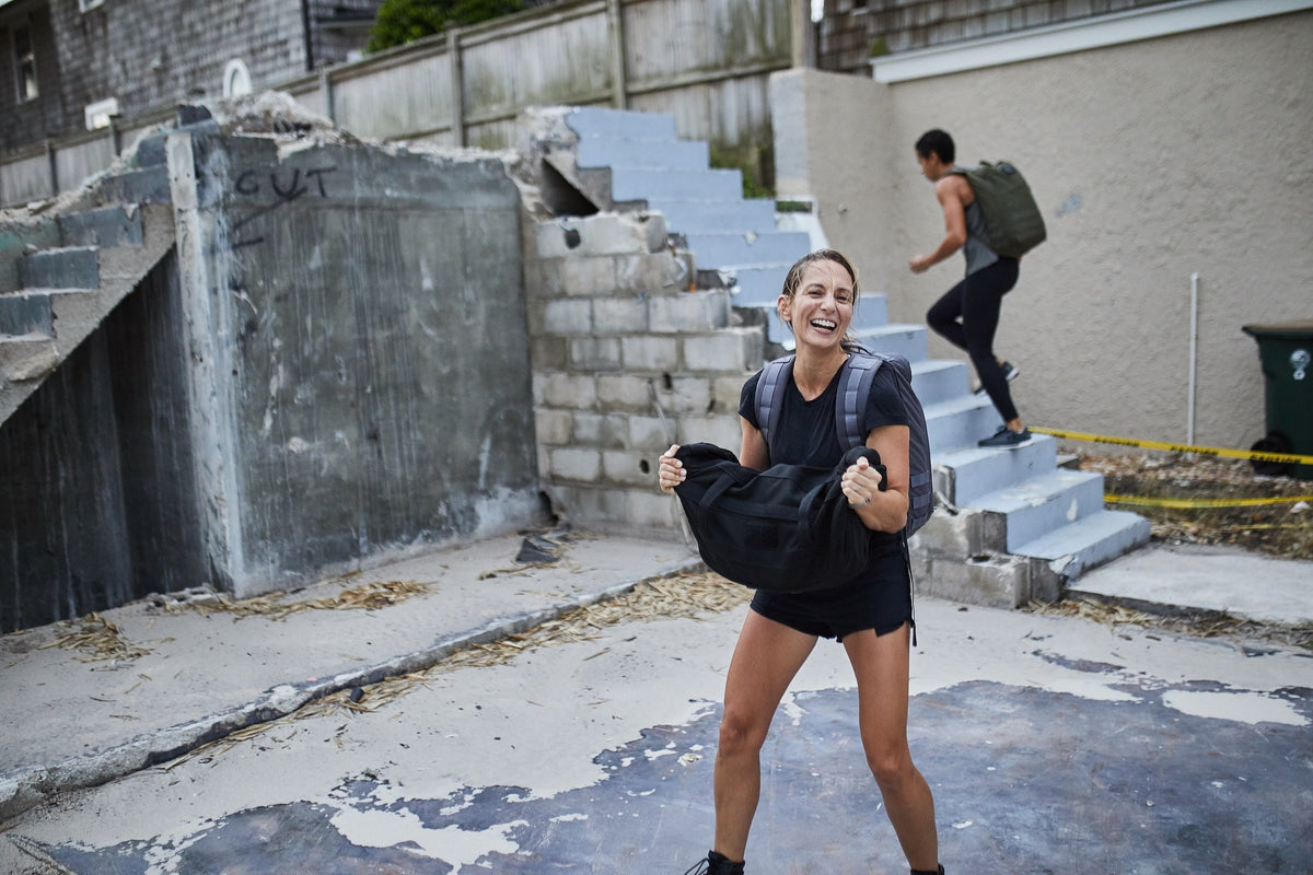Woman training outdoors with GORUCK backpack and sandbag near concrete steps