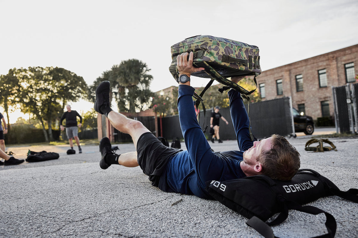 Man performing flutter kicks with camo GORUCK backpack outdoors on pavement during rucking workout